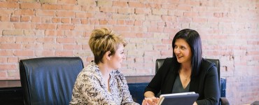 Zwei Frauen an einem Konferenztisch mit Tablet in der Hand, sie unterhalten sich