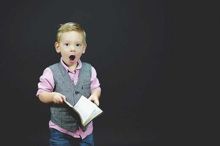 kleiner Junge mit Hemd und Weste, aufgeschlagenes Buch in den Händen, offener Mund